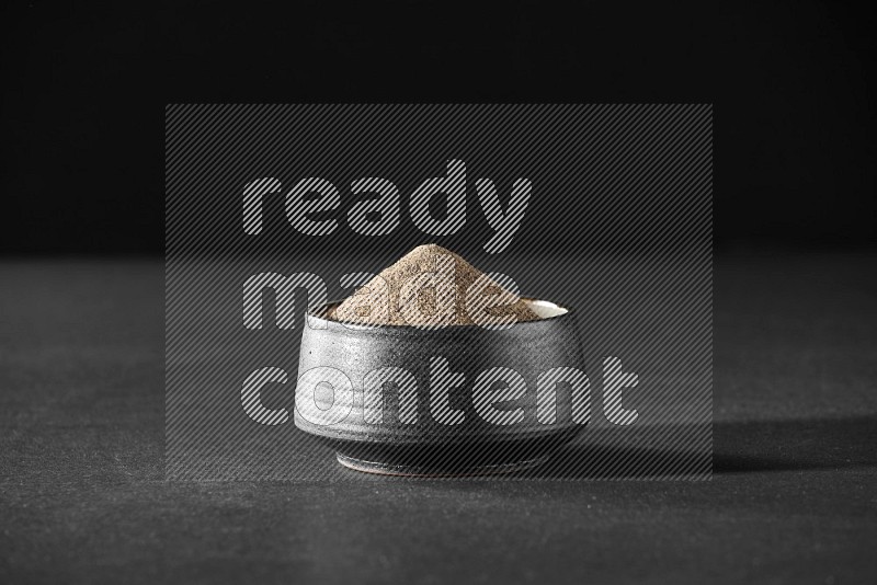 A black pottery bowl full of black pepper powder on black flooring