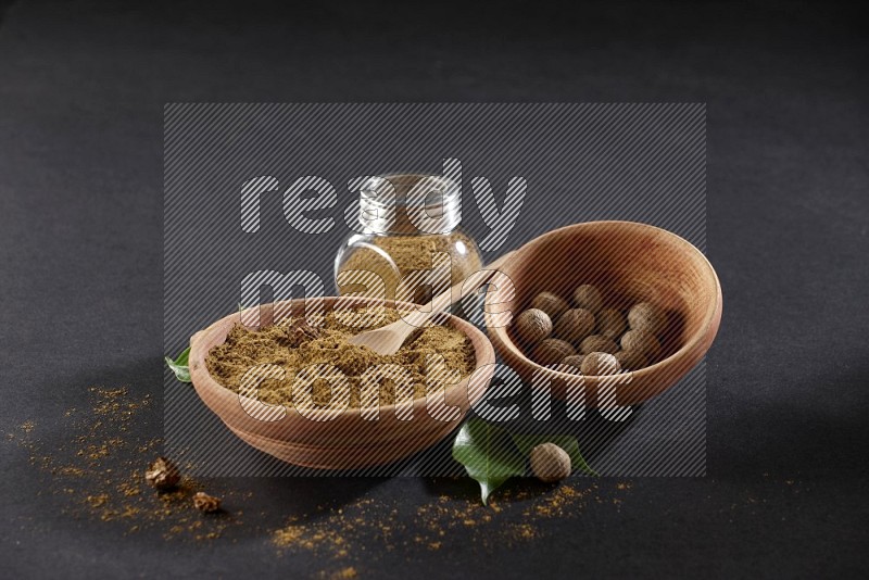 A wooden bowl, spoon and glass spice jar full of nutmeg powder and a wooden bowl full of nutmeg seeds on a black flooring in different angles