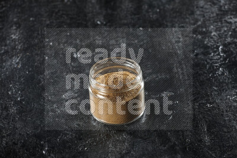 A glass jar full of cumin powder on a textured black flooring