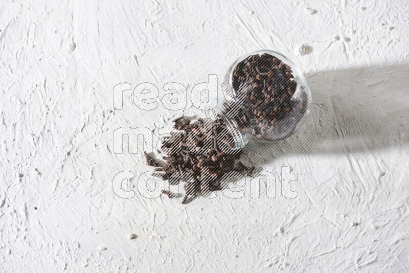 A flipped glass spice jar full of cloves and cloves came out of it on textured white flooring