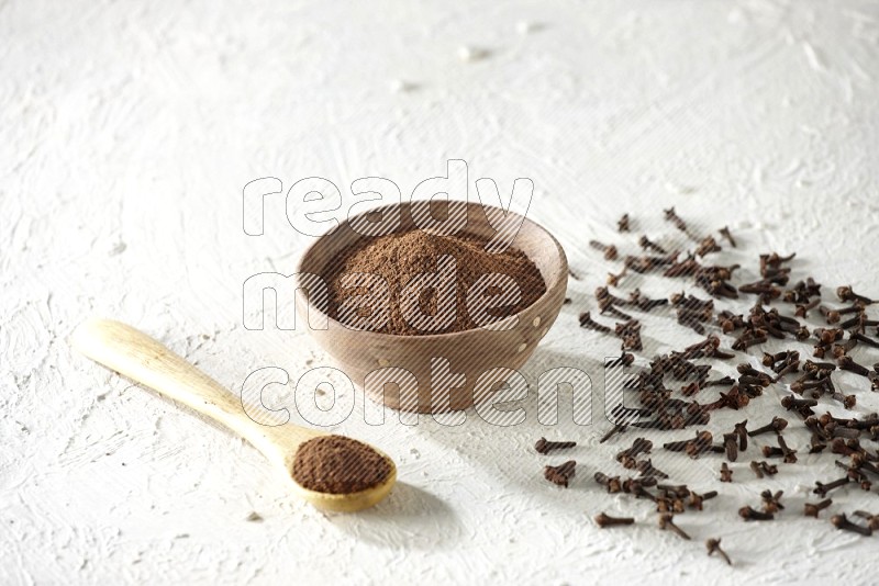 A wooden bowl and wooden spoon full of cloves powder with cloves spread on textured white flooring