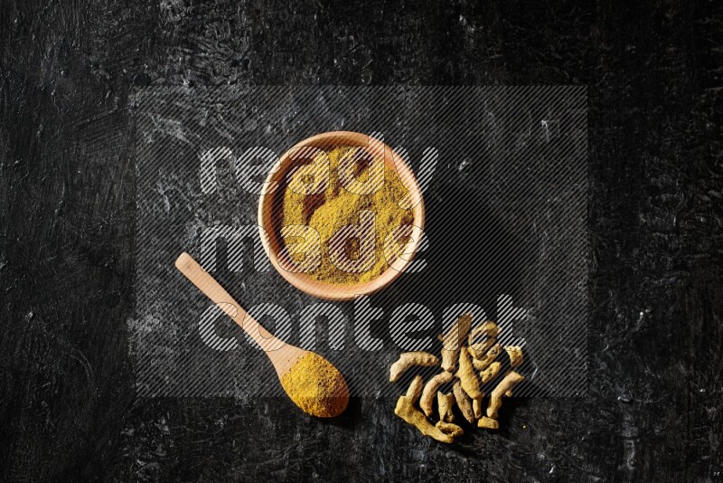 A wooden bowl and a wooden spoon full of turmeric powder with dried turmeric fingers on textured black flooring
