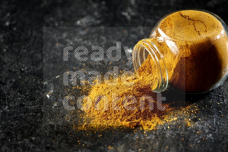 A flipped glass spice jar full of turmeric powder and powder spilled out of it on textured black flooring