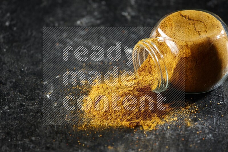 A flipped glass spice jar full of turmeric powder and powder spilled out of it on textured black flooring