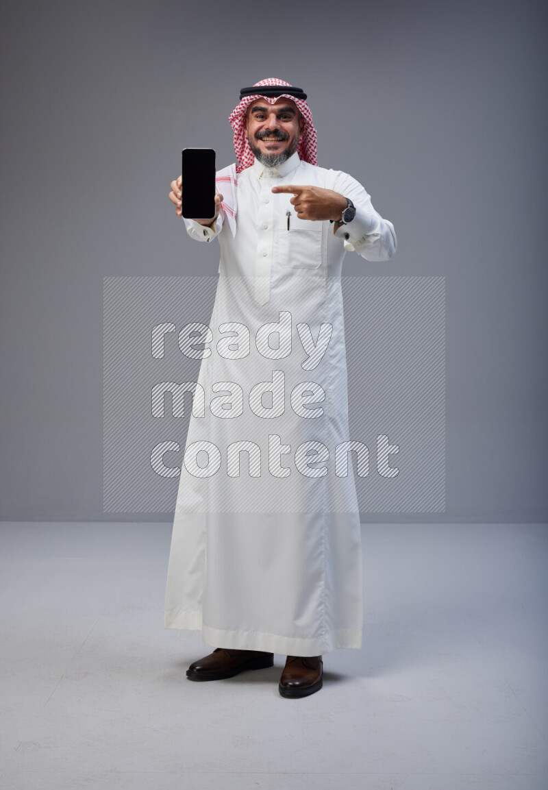 Saudi man Wearing Thob and red Shomag standing showing phone to camera on Gray background