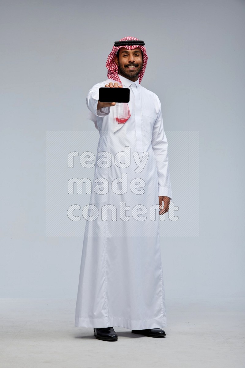 Saudi man Wearing Thob and shomag standing showing phone to camera on Gray background