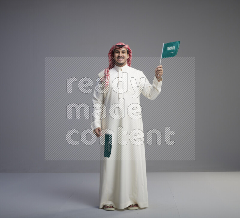 A Saudi man standing wearing thob and red shomag raising small Saudi flag on gray background