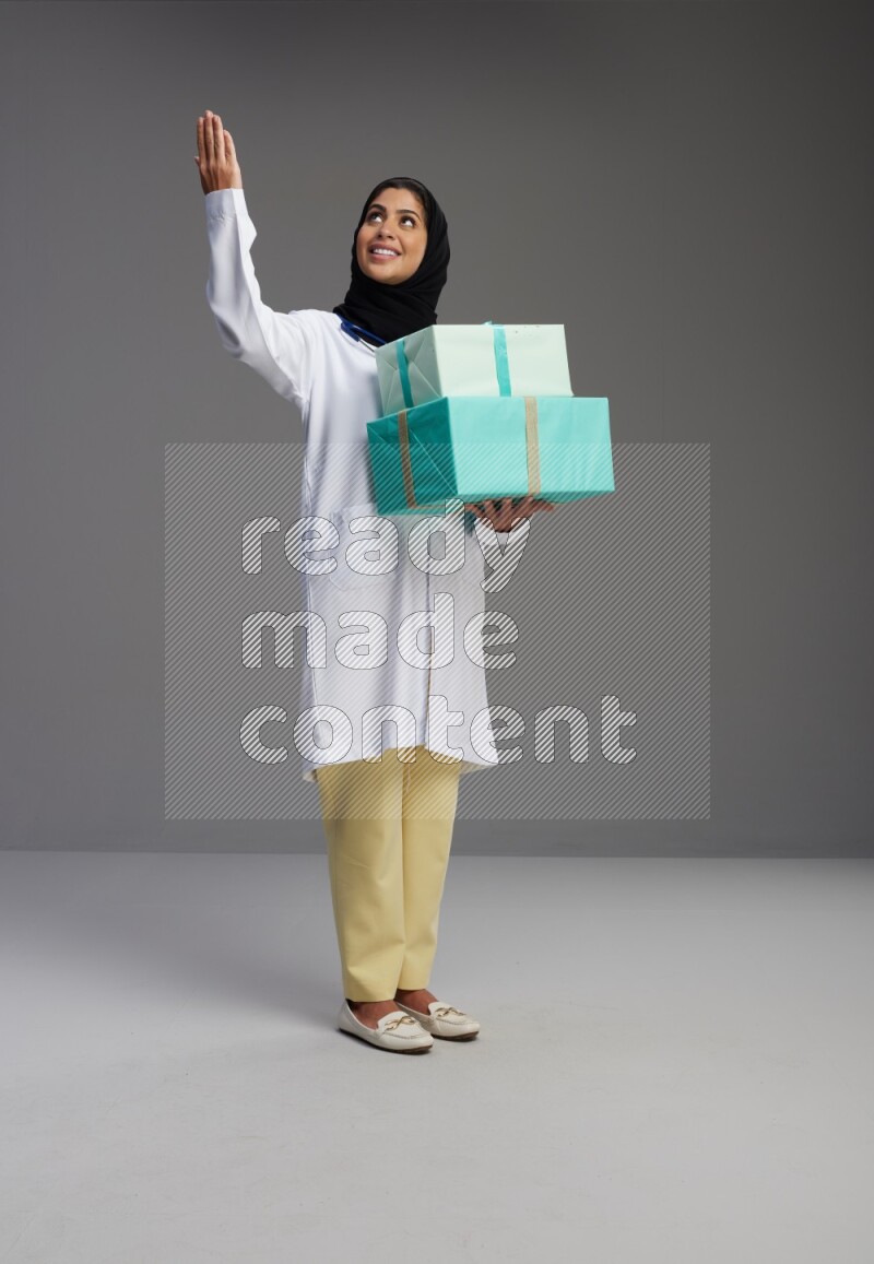 Saudi woman wearing lab coat with stethoscope standing holding gift box on Gray background