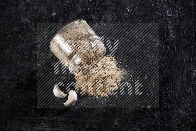 A glass jar full of garlic powder flipped over with the powder came out on a textured black flooring