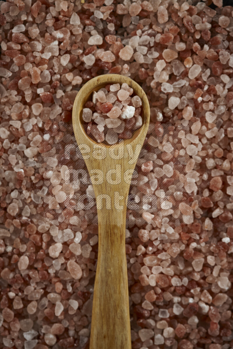 A wooden spoon full of coarse himalayan salt crystals on a bunch of the crystals on black background