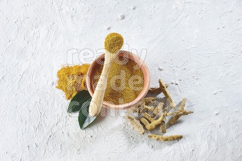 A wooden bowl and wooden spoon full of turmeric powder with dried turmeric fingers beside it on textured white flooring