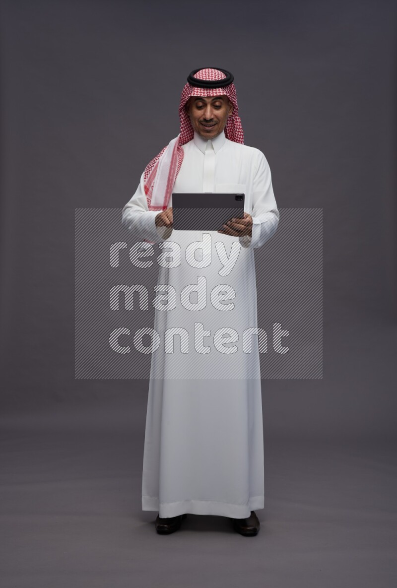 Saudi man wearing thob and shomag standing working on tablet on gray background