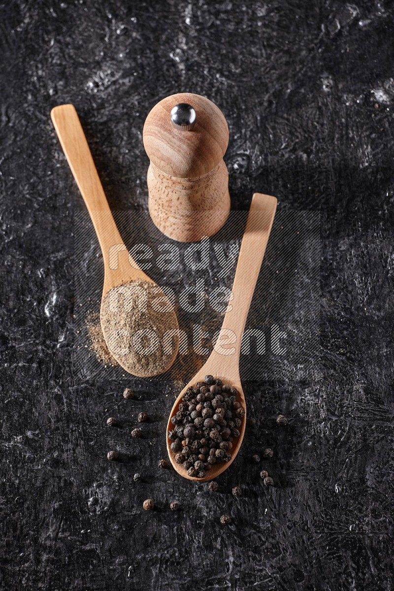 Black pepper powder and black pepper beads in wooden spoons and wooden grinder on a textured black flooring