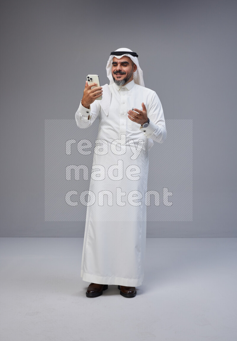 Saudi man Wearing Thob and white Shomag standing texting on phone on Gray background