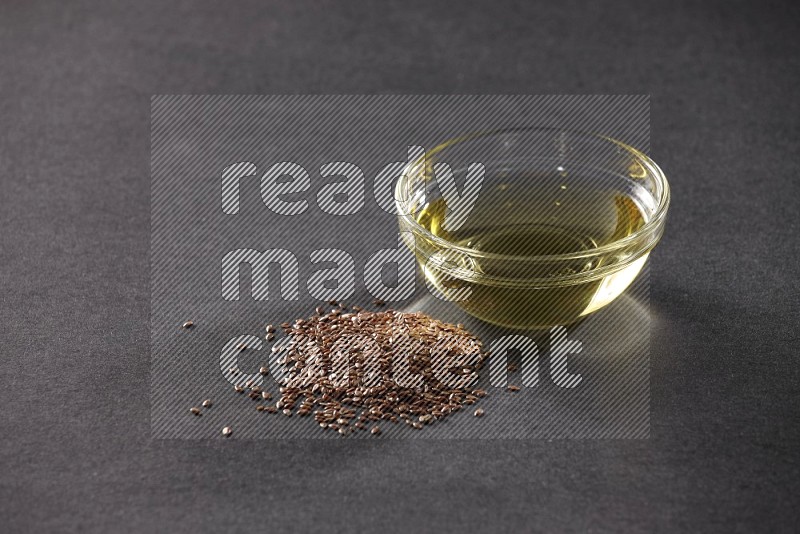 A glass bowl full of flaxseeds oil with some of flaxseeds beside it on a black flooring