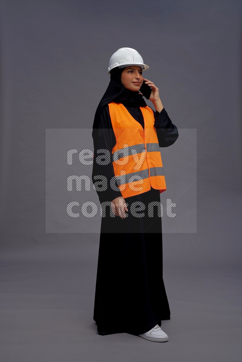 Saudi woman wearing Abaya with engineer vest standing talking on phone on gray background