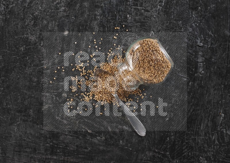 A glass spice jar and a metal spoon full of mustard seeds and jar is flipped with fallen seeds on a textured black flooring