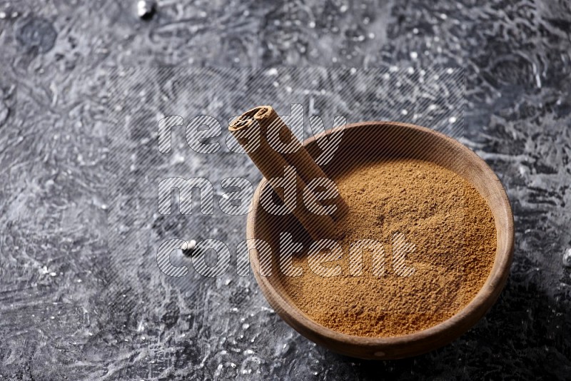 Wooden bowl full of cinnamon powder and a cinnamon stick on a textured black background