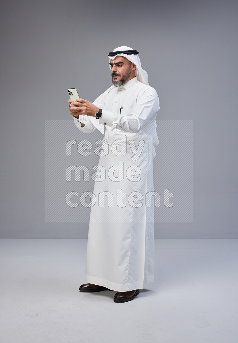 Saudi man Wearing Thob and white Shomag standing texting on phone on Gray background