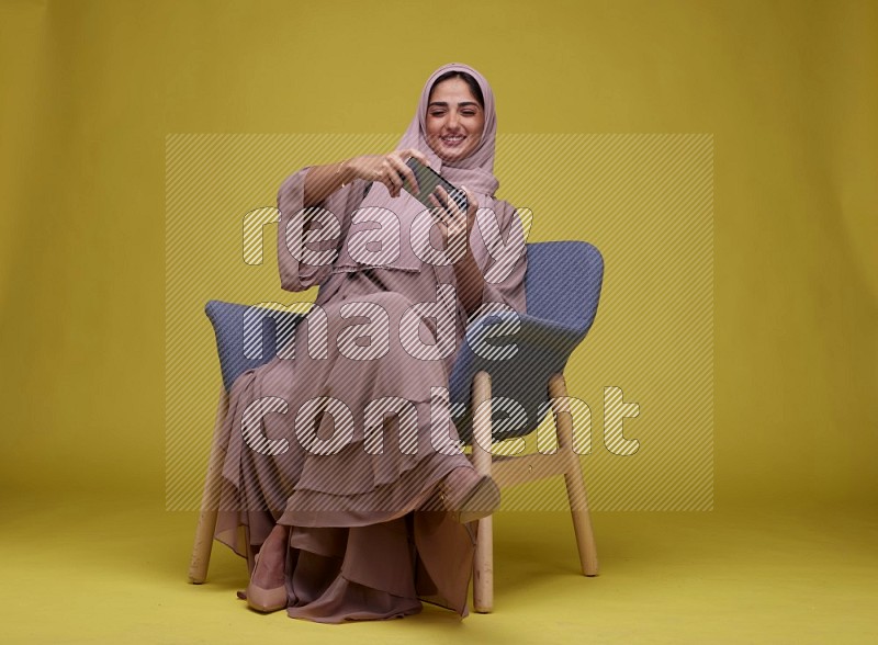 A woman Playing Game Sitting  on a Yellow Background wearing Brown Abaya with Hijab