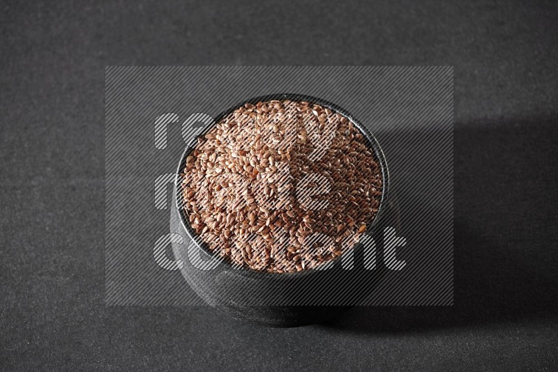 A black pottery bowl full of flaxseeds on a black flooring