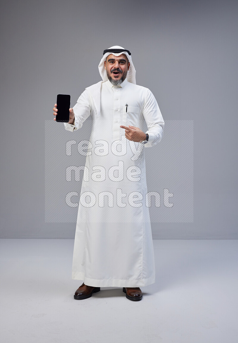 Saudi man Wearing Thob and white Shomag standing showing phone to camera on Gray background