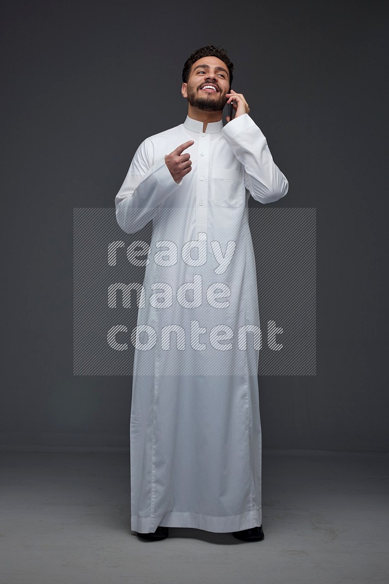 A Saudi man wearing Thobe and talking in the phone while standing and making different poses eye level on a gray background