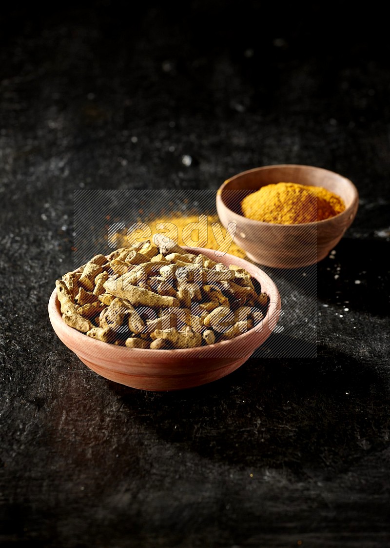 2 wooden bowls, one full of turmeric powder and the other full of dried turmeric whole fingers on a textured black flooring