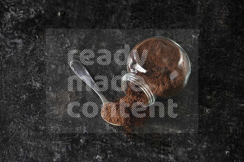 A flipped glass spice jar and a metal spoon full of cloves powder on textured black flooring