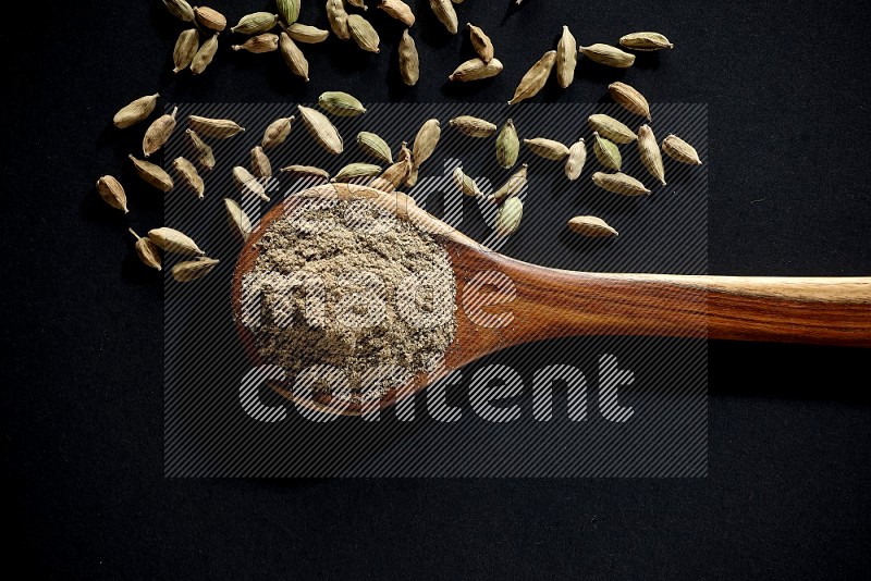 A wooden ladle full of cardamom powder and cardamom seeds beside it on black flooring