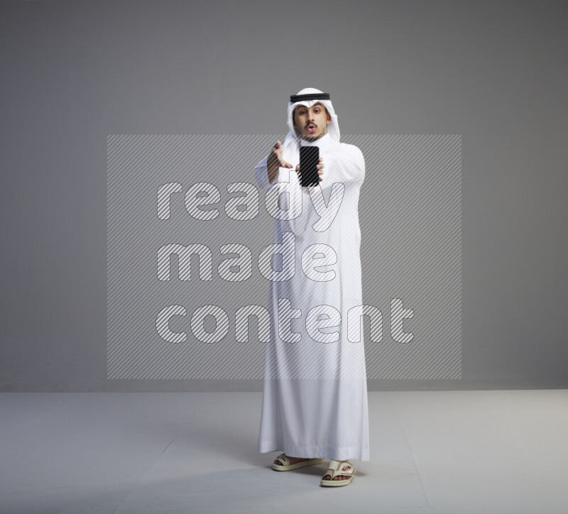 A Saudi man standing wearing thob and white shomag showing phone to camera on gray background