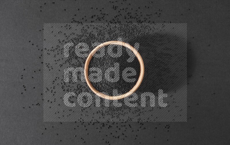 A wooden bowl full of black seeds surrounded by seeds on a black flooring