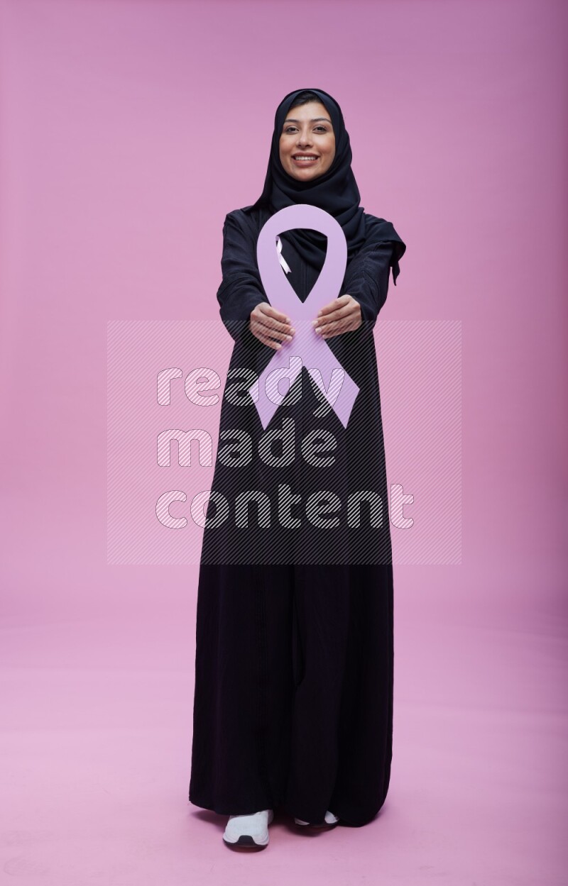 Saudi woman wearing pink ribbon on abaya standing holding awareness ribbon on plain pink background
