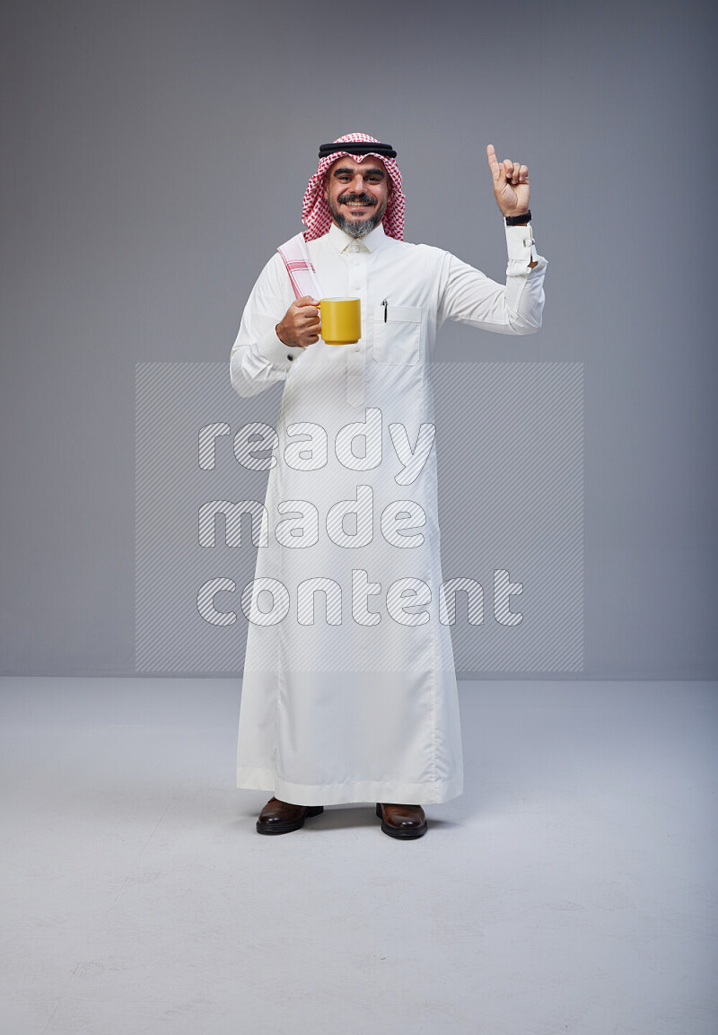 Saudi man Wearing Thob and red Shomag standing holding mug on Gray background