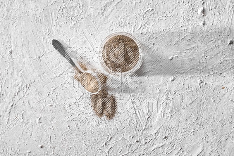 A glass jar full of black pepper powder and a metal spoon full of powder on a textured white flooring