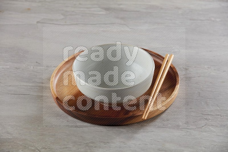 white pottery round bowl on top of brown wood round plate and wood chopsticks, on grey textured countertop