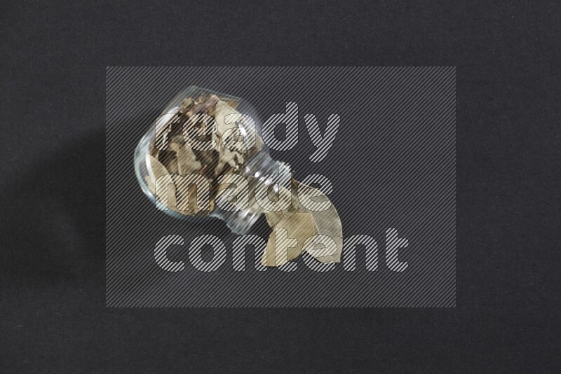 A glass spice jar full of dried bay leaves and jar is flipped and leaves spread out on black flooring