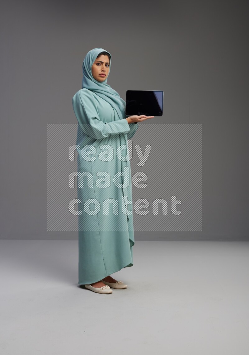 Saudi Woman wearing Abaya standing showing tablet to camera on Gray background