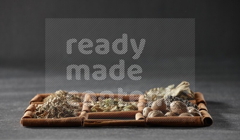 9 مربعات من أعواد القرفة ممتلئة بالهيل في المنتصف محاطة بجوزة الطيب والفلفل الأسود وورق الغار والبهارات والكمون والفلفل الأبيض والريحان المجفف والملح على أرضية سوداء