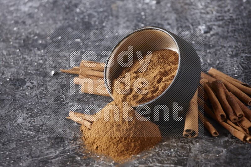 Black pottery bowl over filled with cinnamon powder and cinnamon sticks around the bowl on a textured black background