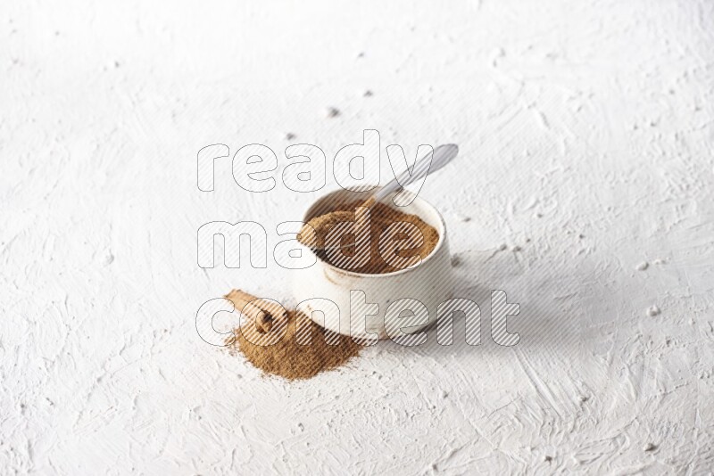 Ceramic beige bowl full of cinnamon powder and a metal spoon with cinnamon sticks next of it on a textured white background
