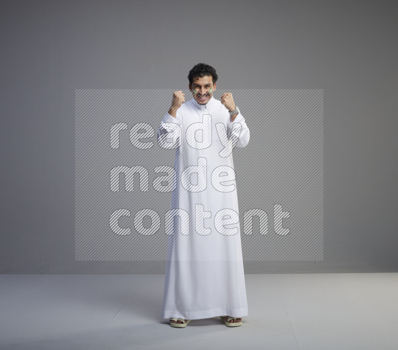 A Saudi man standing wearing thob with face painting interacting with the camera on gray background