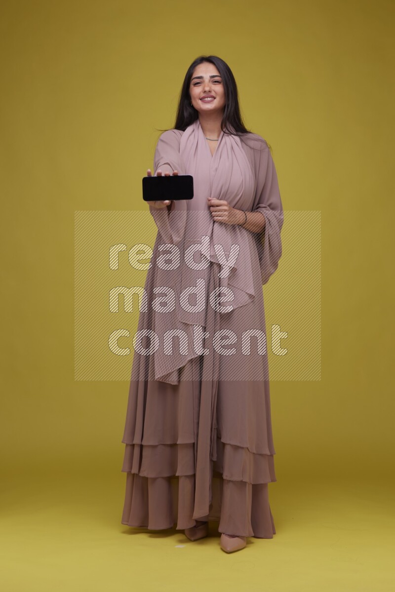 A woman Showing SmartPhone Screen on a Yellow Background wearing Brown Abaya