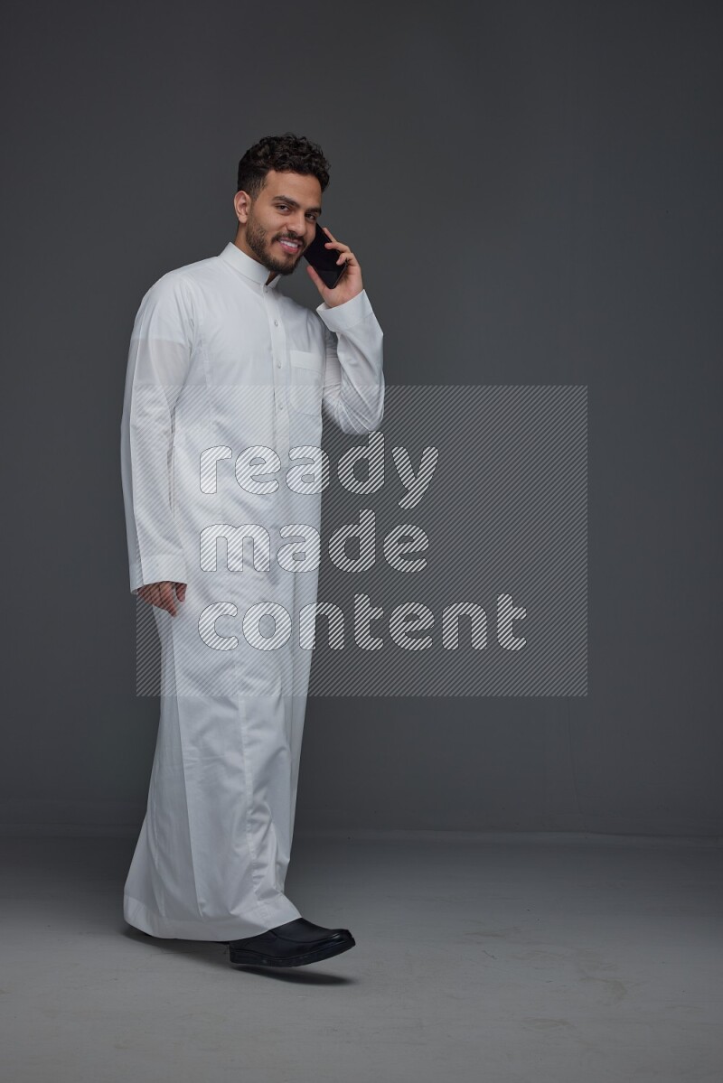 A Saudi man wearing Thobe and talking in the phone while standing and making different poses eye level on a gray background