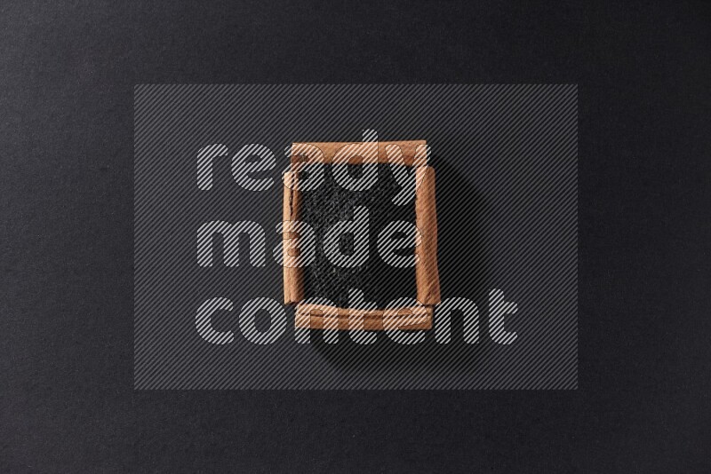 أربعة من أعواد القرفة مكونة شكل مربع بداخلها حبة البركة علي خلفية سوداء