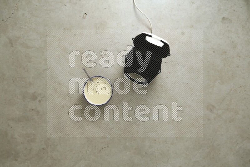 A ceramic bowl beside a carton of eggs and glass bowl full of sugar, vanilla extract, flour on beige marble background