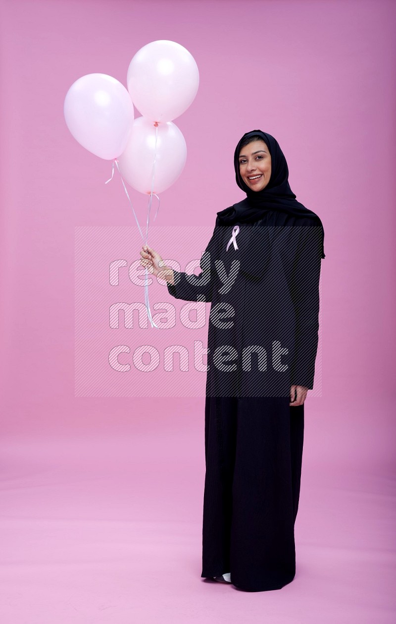 Saudi woman wearing pink ribbon on Abaya standing holding balloon on plain pink background