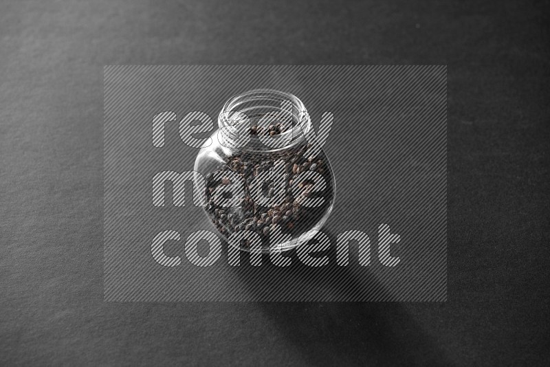 A glass spice jar full of black peppers on black flooring