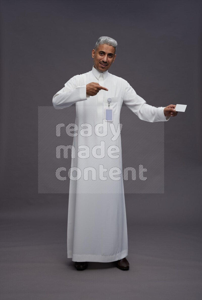 Saudi man wearing thob with pocket employee badge standing holding ATM card on gray background