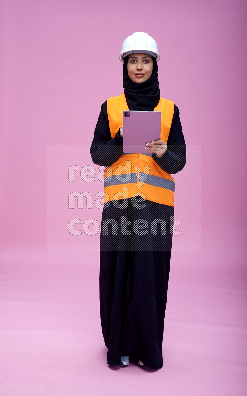 Saudi woman wearing Abaya with engineer vest and helmet standing working on tablet on pink background